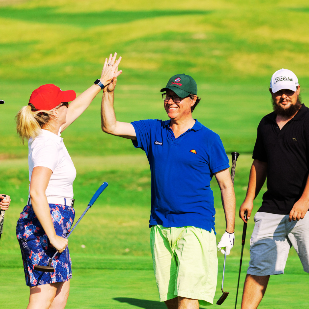 Two golfers share a high-five on the fairway, smiling as teammates look on.